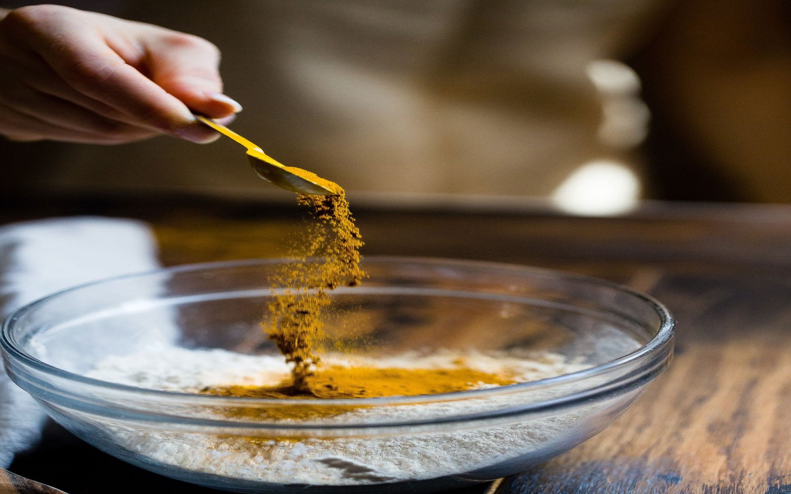 A hand (with spoon) pouring the turmeric in a bowl with some substance in it.